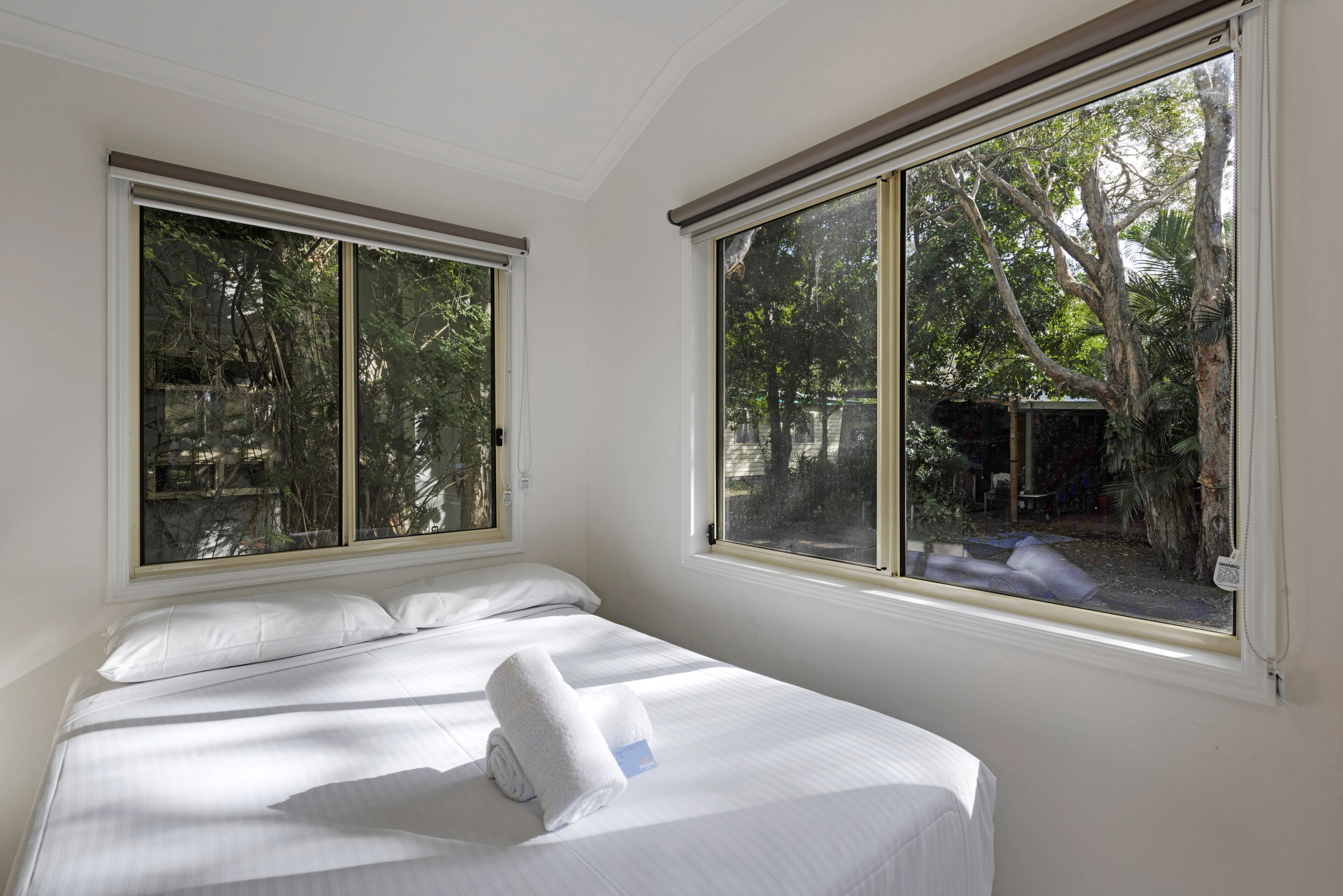 A neatly made bed with a folded towel, large windows with blinds, and a view of trees and a fence outside.