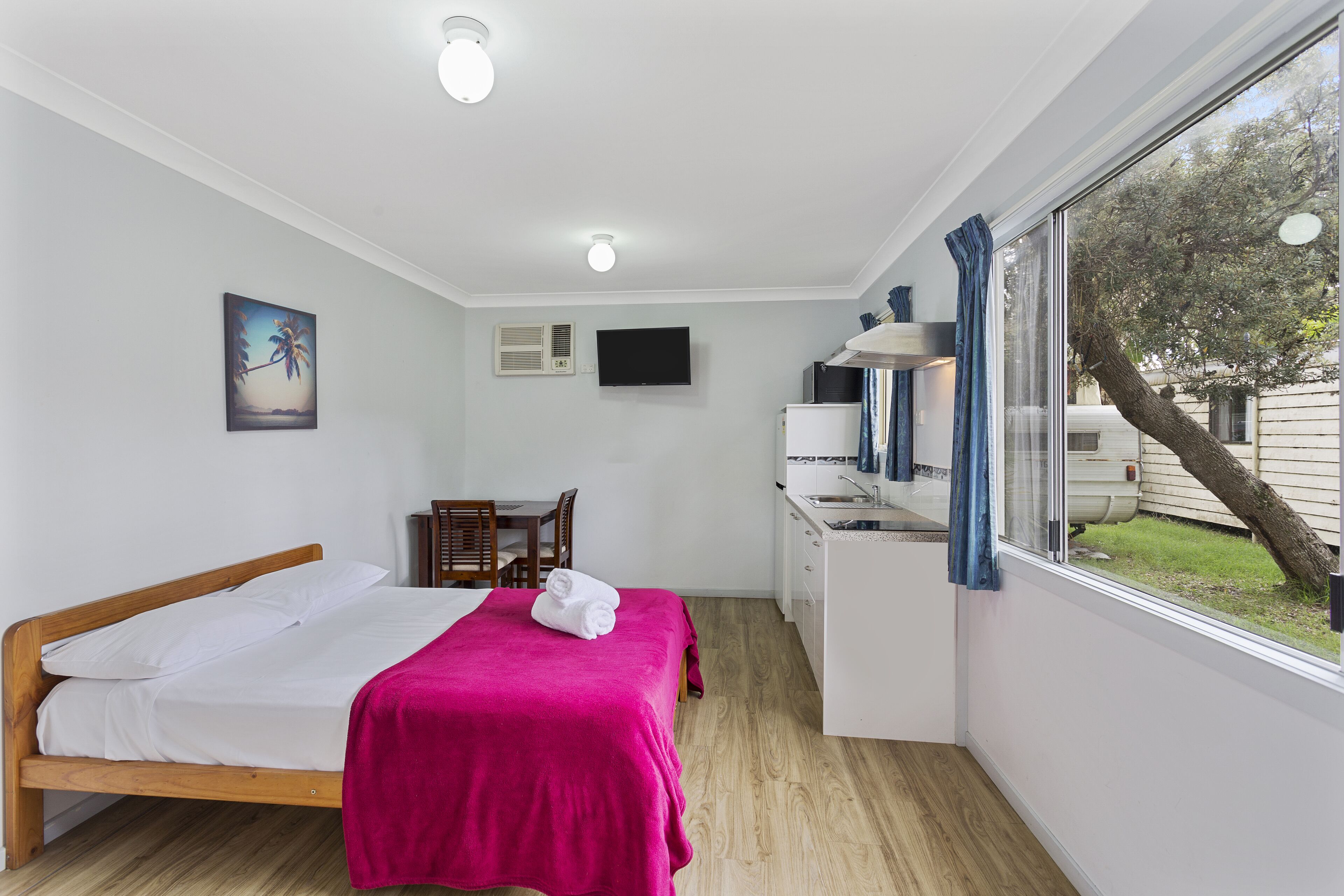 A compact room with a bed, a desk, a chair, a TV, and a window with a view of the outdoors.