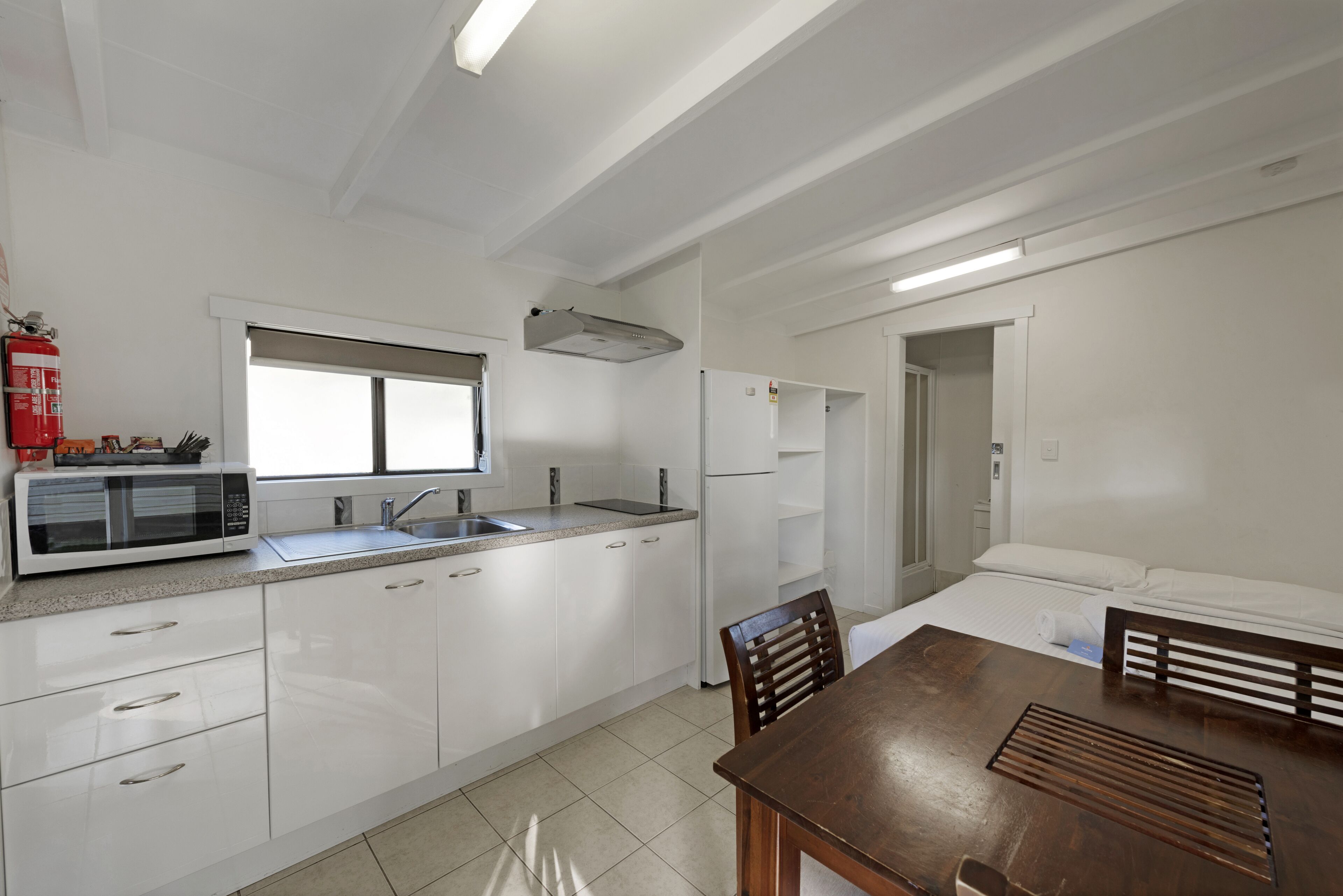 Una cocina moderna con armarios blancos, un microondas y un fregadero. Hay un comedor con una mesa y sillas de madera.