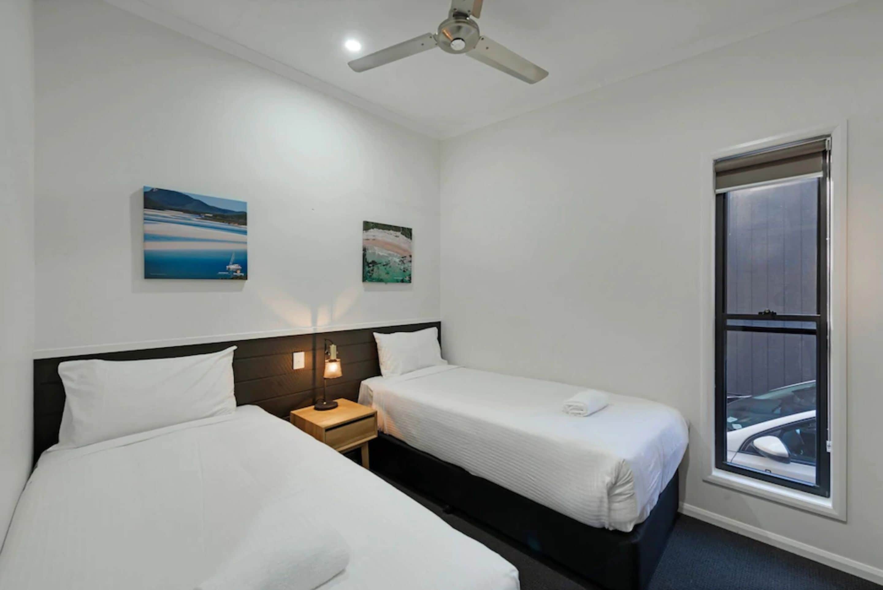 A hotel room with two single beds, a ceiling fan, a window with a view of a car, and two wall-mounted paintings.