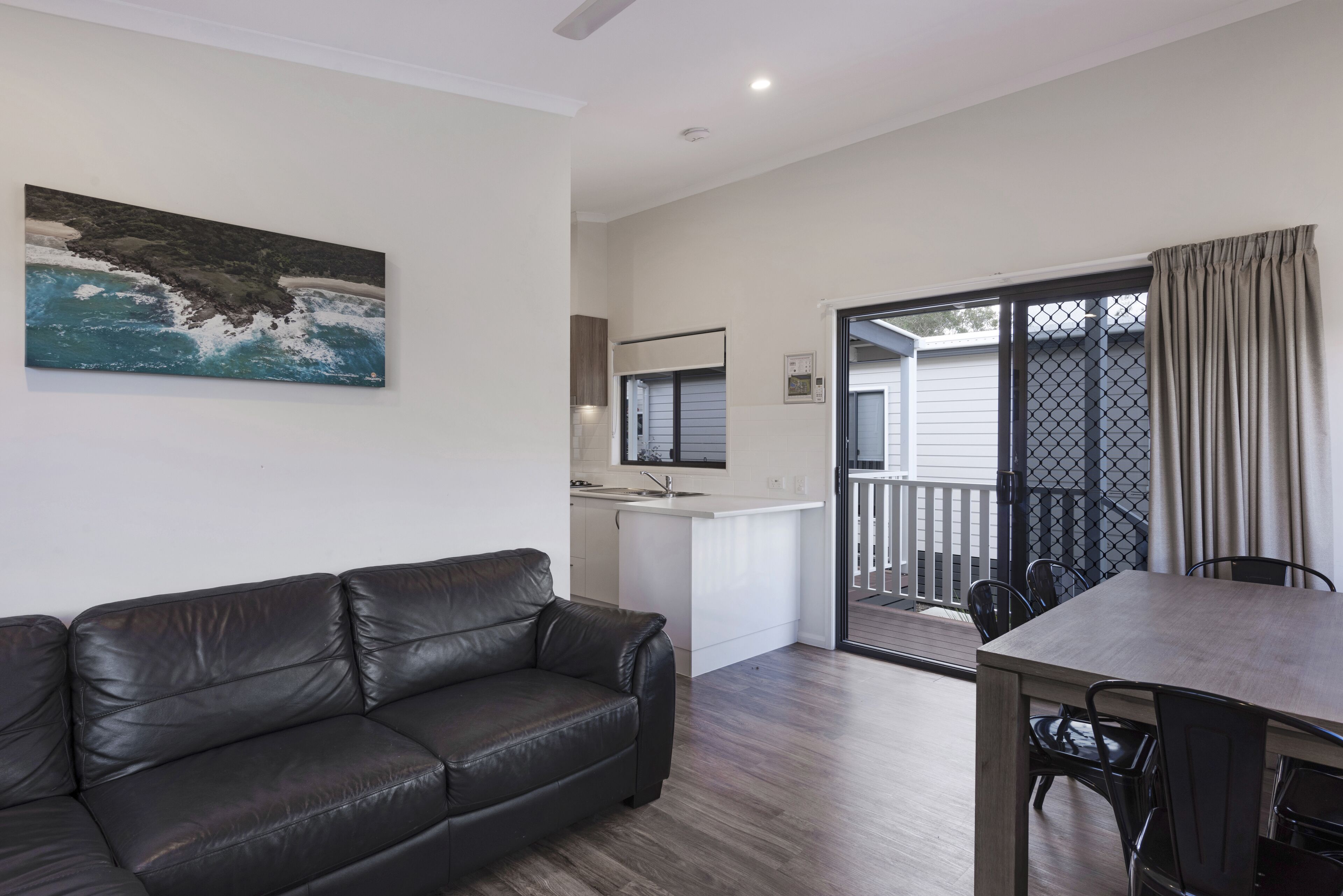 Un salon moderne avec un canapé en cuir noir, une table à manger avec des chaises, un coin cuisine et un balcon offrant une vue sur l’extérieur.
