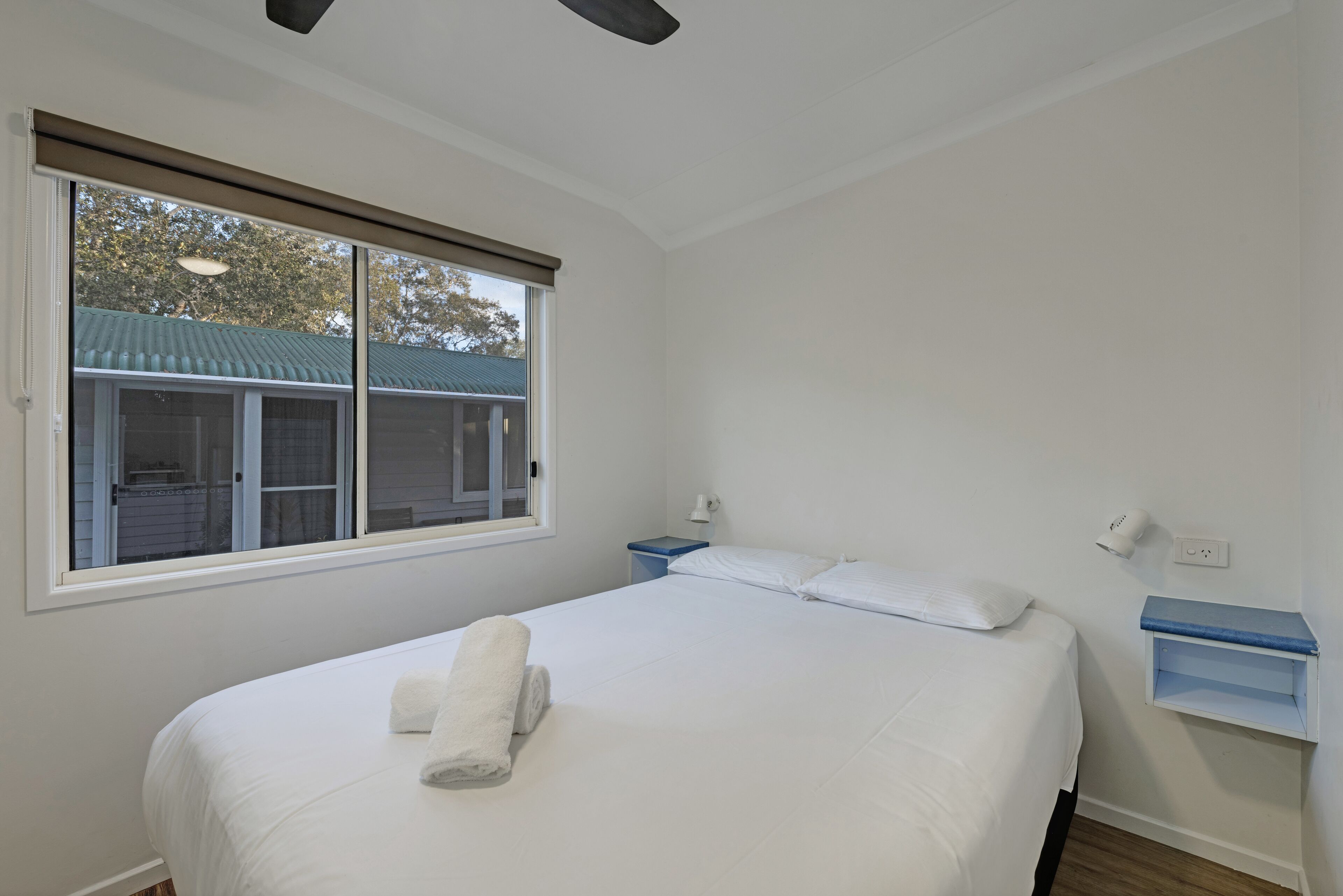 A bedroom with a large bed, a window with blinds, a bedside table, and a nightstand.