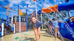 Zona de juegos acuáticos con toboganes y chorros de agua, que incluye una pared de ladrillo y un tobogán de color azul y naranja.
