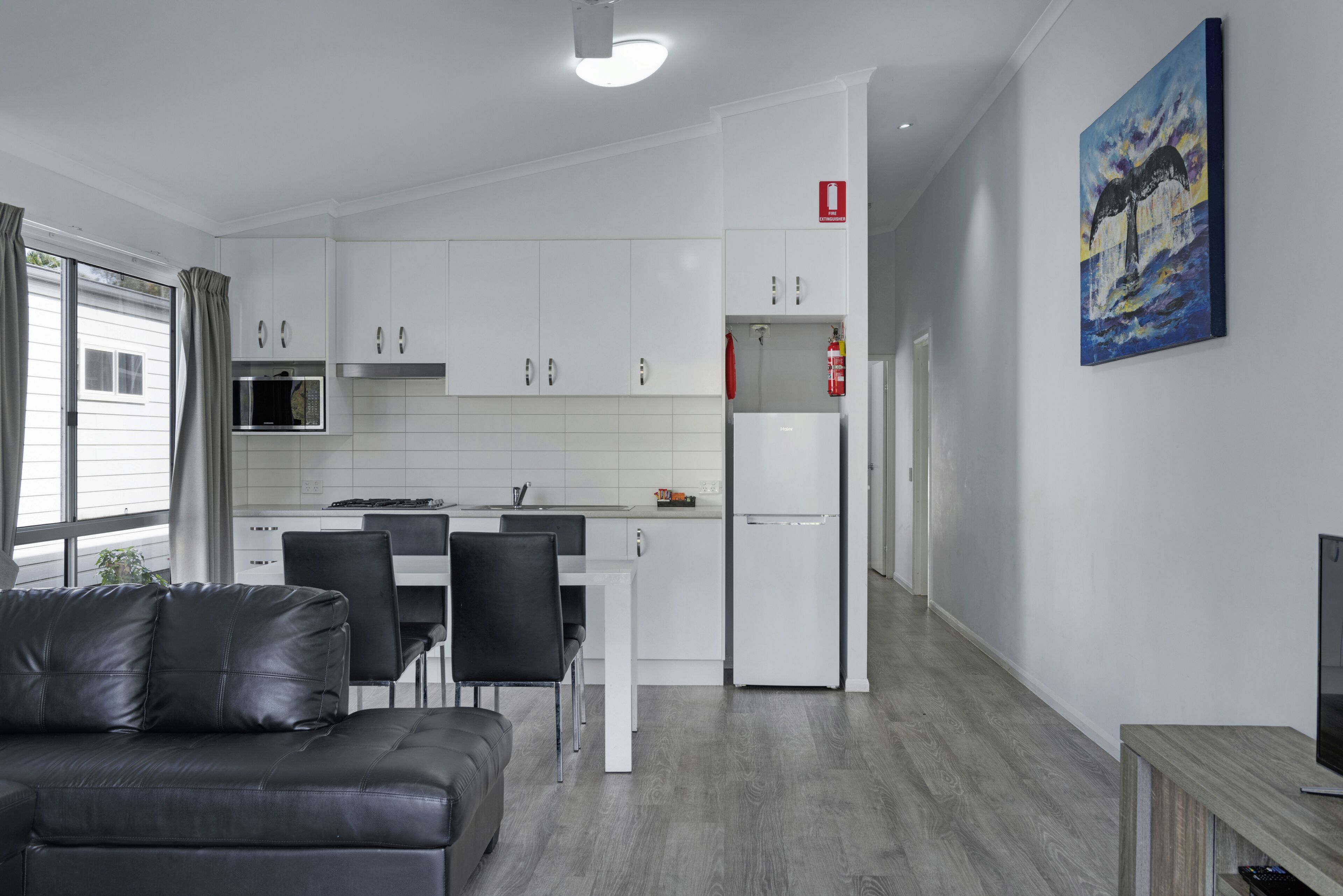 A modern kitchen with white cabinets, a microwave, and a refrigerator. There is a dining table with chairs, a black leather sofa, and a painting on the wall.