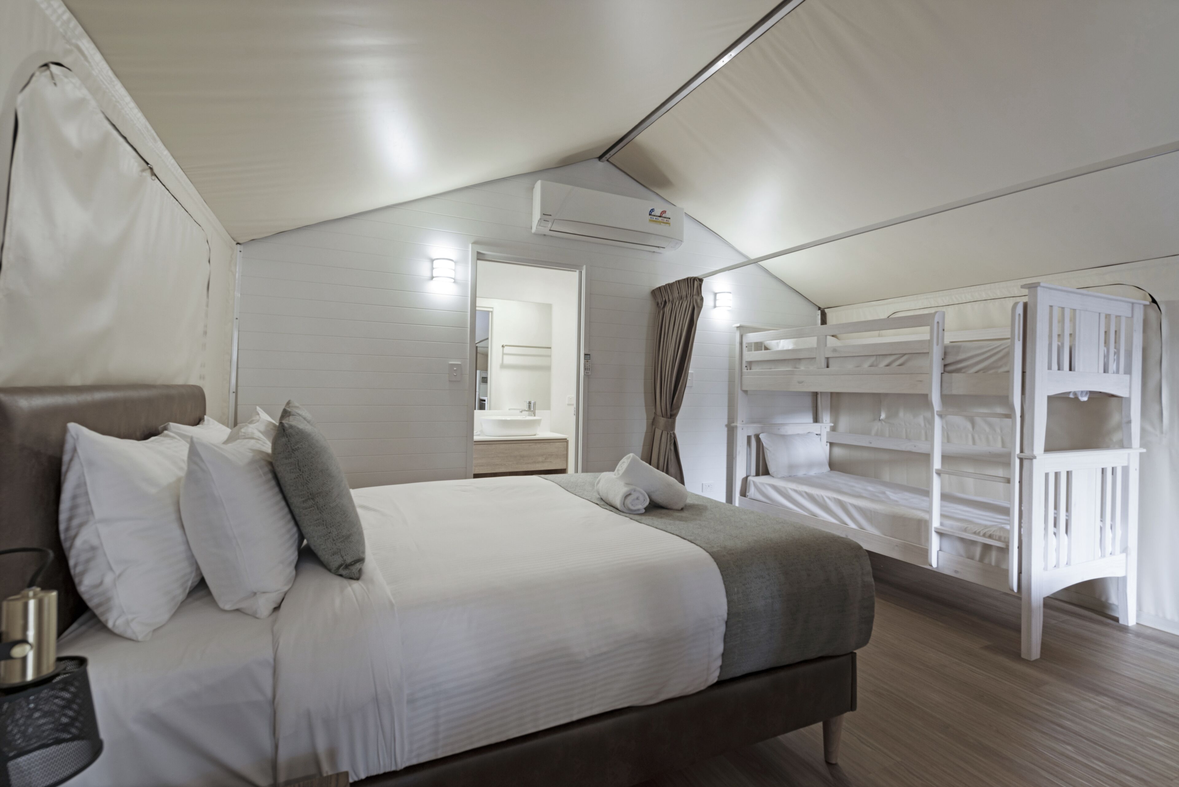 A bedroom with a bed, pillows, a bedside table, a bunk bed, and an air conditioning unit.