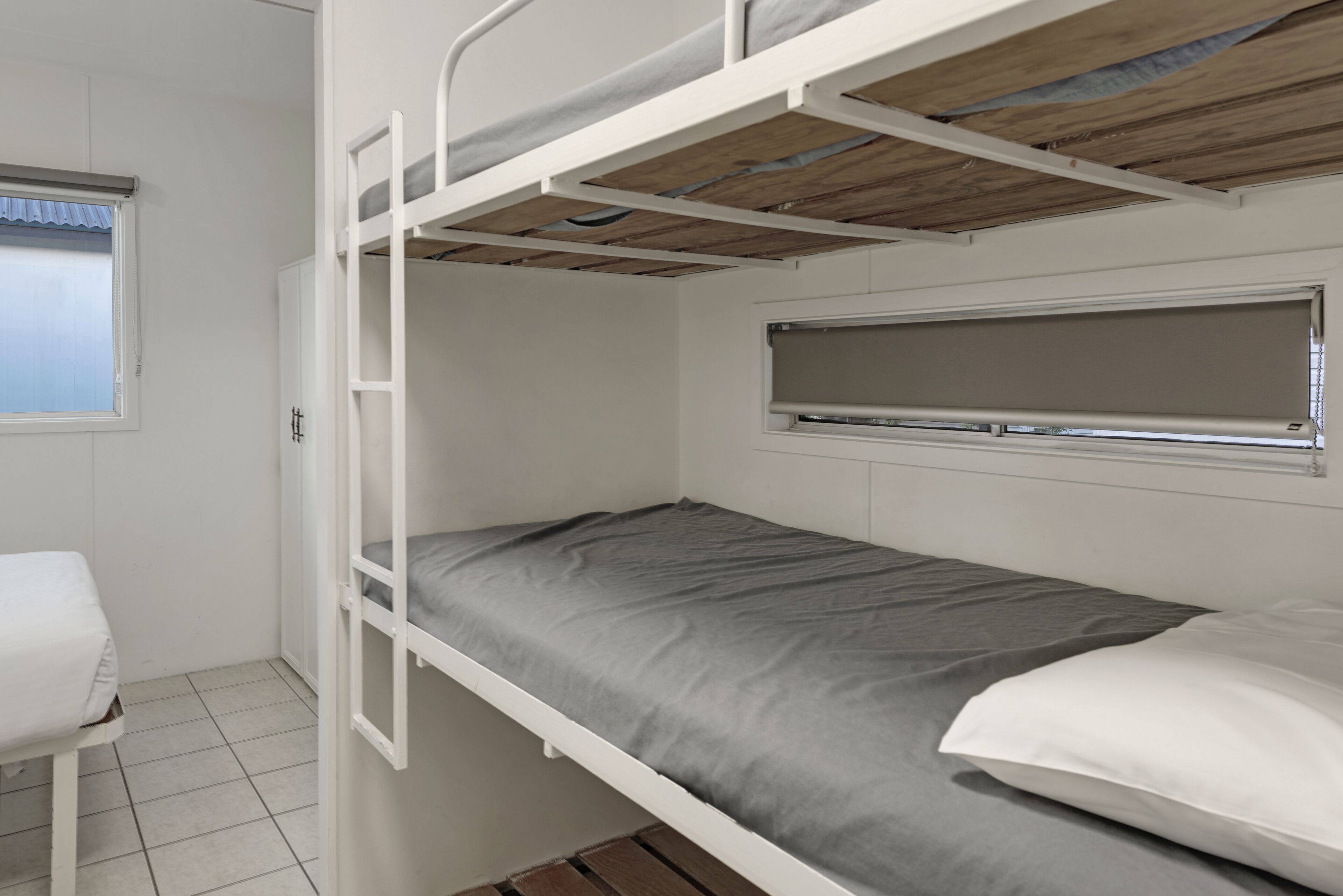 A bunk bed with a grey mattress and white pillows, a window with blinds, and a white wardrobe.
