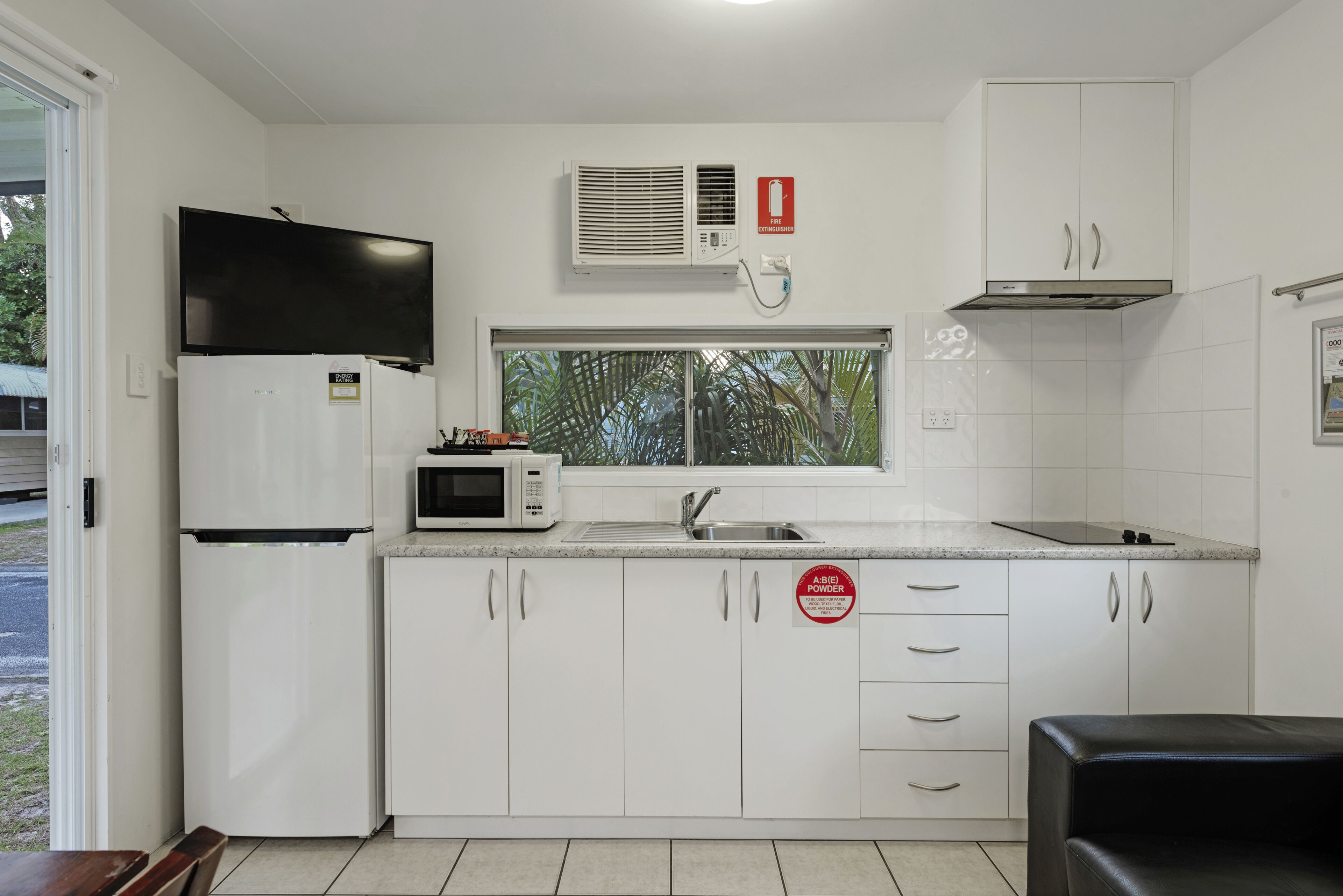 A compact kitchen with white cabinets, a microwave, a refrigerator, a sink, and a window with a view of trees.