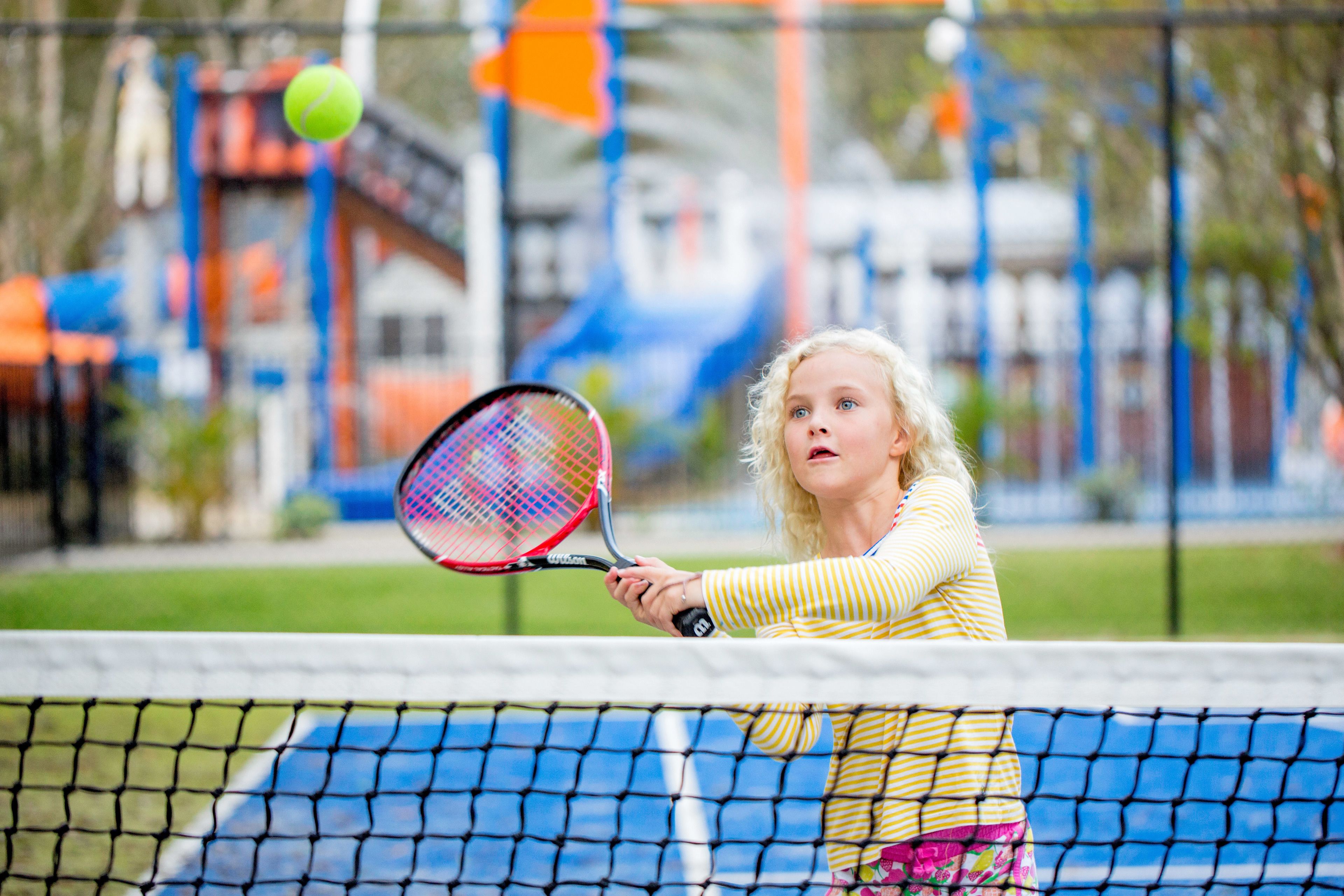 A tennis court with a net in the foreground and a colorful playground structure in the background.