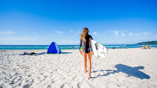 Escena de playa con una tienda de campaña y una persona llevando una tabla de surf.