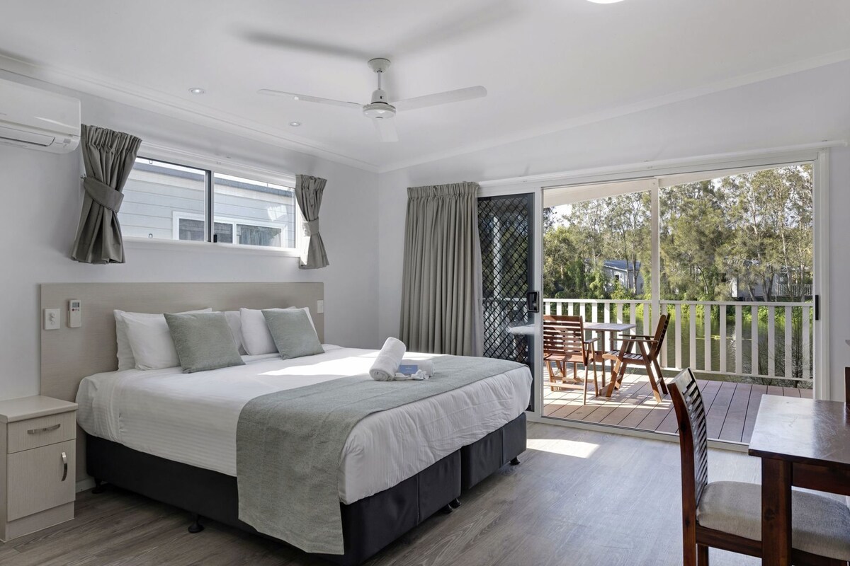 Quarto com cama, mesa de cabeceira e vista para uma varanda com mobiliário exterior.