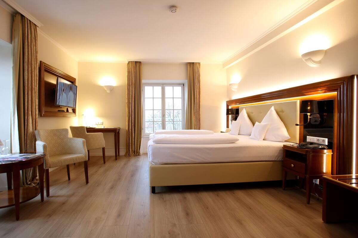 Una habitación de hotel con una cama grande, dos mesitas de noche, un escritorio con una silla, un televisor en la pared y una ventana con cortinas.