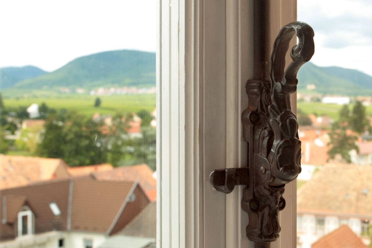Una ventana con un tirador metálico ornamentado y vistas a un pueblo con edificios de techo rojo y colinas verdes al fondo.