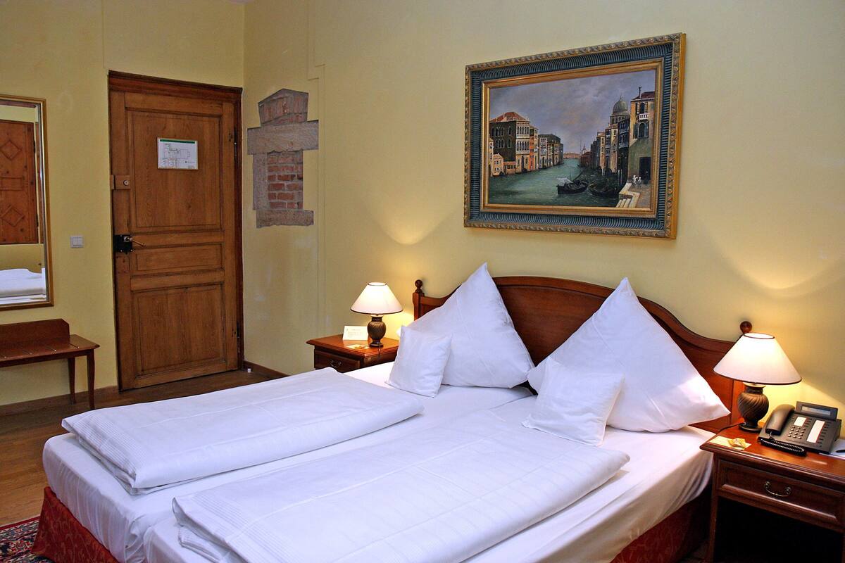 Habitación de hotel con cama doble, dos mesitas de noche con lámparas, cabecera de madera, un cuadro de un paisaje urbano y una puerta con un cartel.