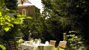 Un área de comedor al aire libre con una mesa lista para dos personas, rodeada de follaje y estatuas.