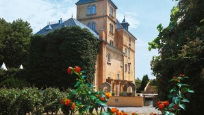 Un edificio grande e histórico con una torre destacada y numerosas ventanas, rodeado de un jardín bien cuidado con plantas florecientes.