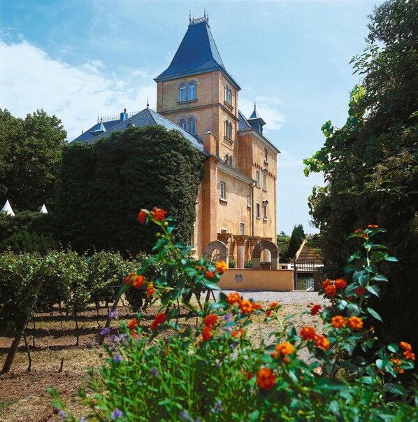 A large, historic building with a prominent tower and multiple windows, surrounded by a well-maintained garden with flowering plants.
