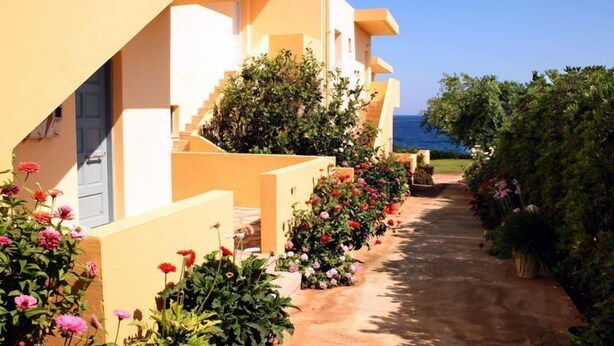 A row of Mediterranean-style buildings with a pathway lined with colorful flowers leading to a body of water. - Nautilus Apartments (Agios Nikolaos)