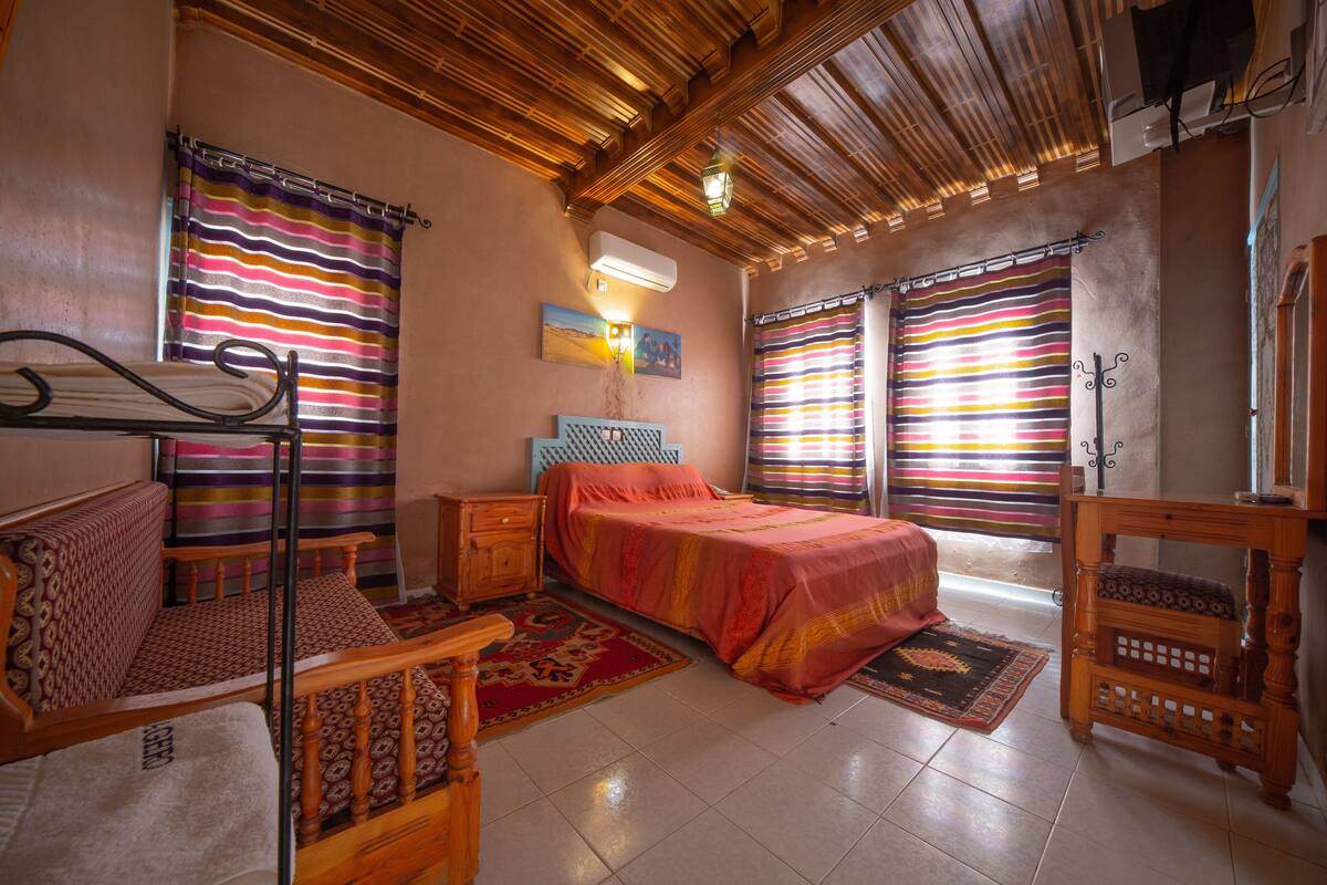 Un dormitorio con una cama, un banco de madera, una mesita de noche, una lámpara y una ventana con cortinas a rayas.