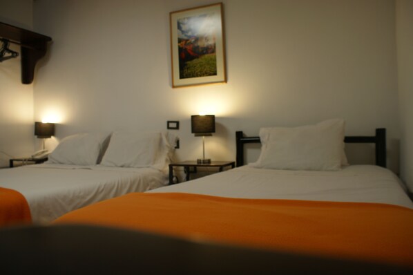 Habitación de hotel con dos camas, cada una con ropa blanca y una manta color naranja. Se observan mesitas de noche con lámparas y un cuadro en la pared.