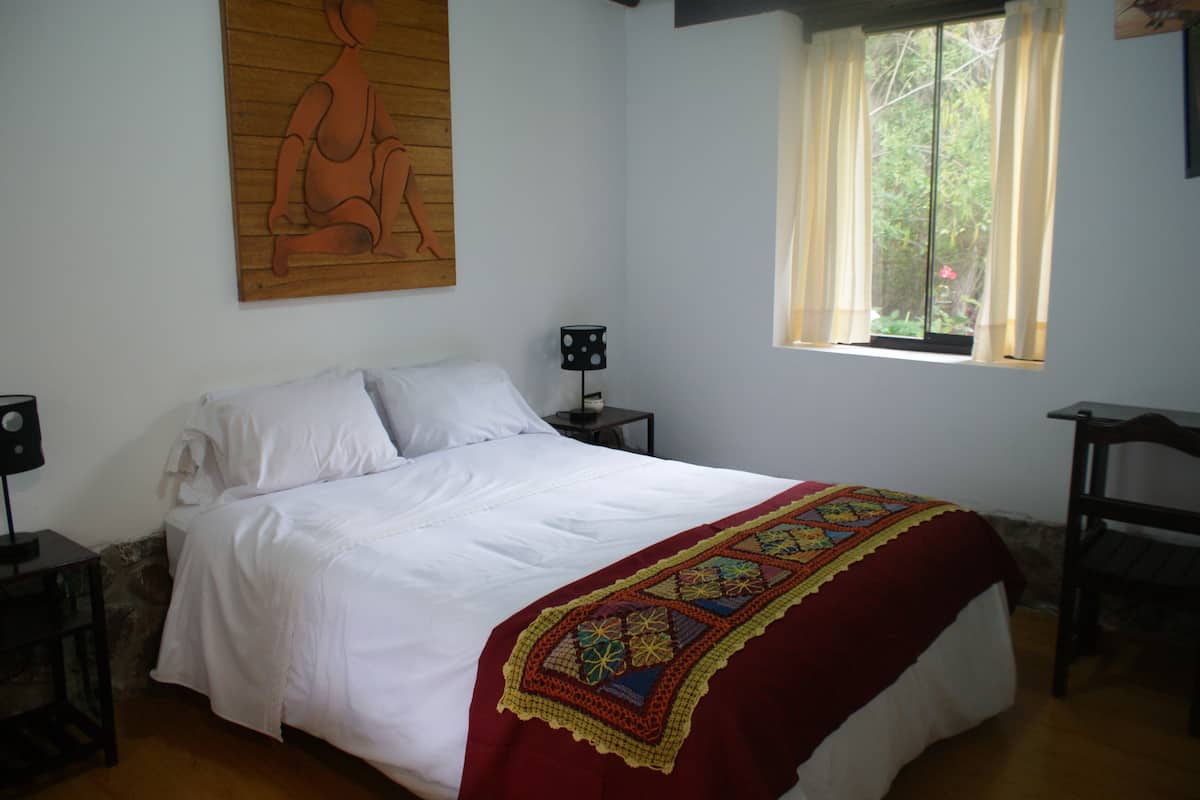 A bedroom with a bed, a window with curtains, a nightstand, and a wall-mounted painting.