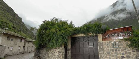 Un edificio de paredes de piedra con una verja metálica cerrada, una calle empedrada y montañas al fondo.