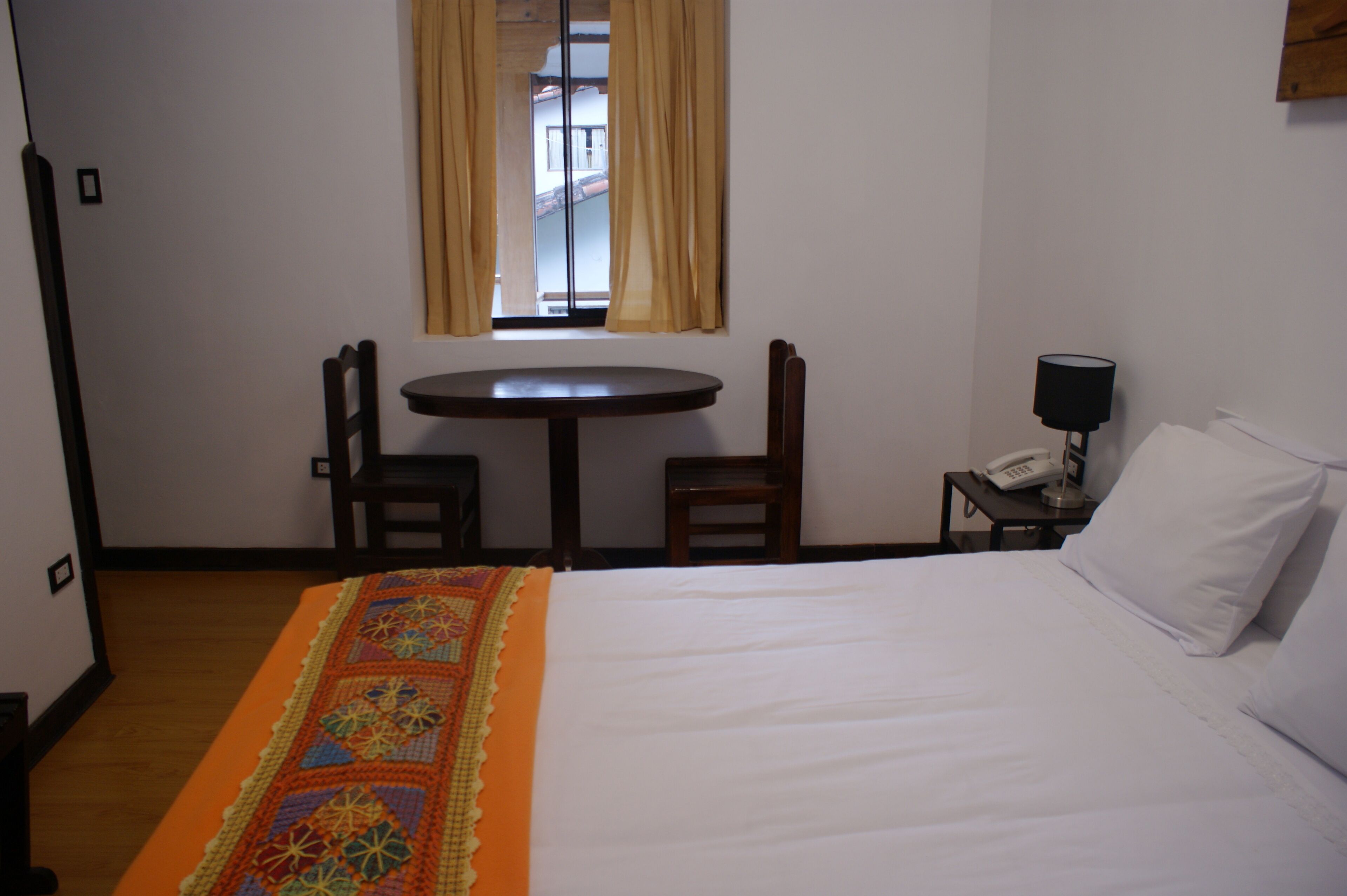 A hotel room with a bed, a small table, two chairs, a nightstand with a lamp, and a window with curtains.
