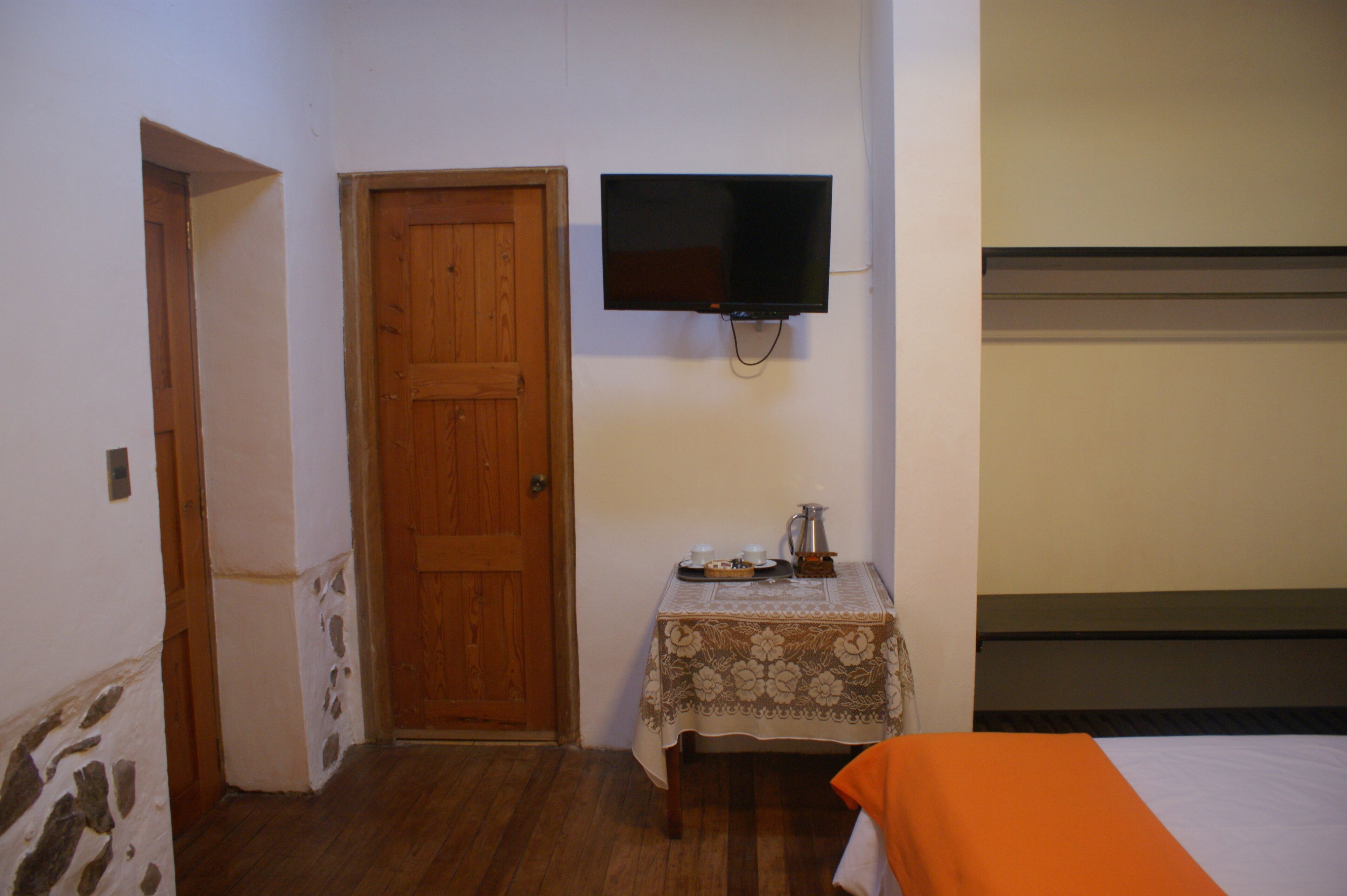 Ein Zimmer mit Holztür, einem an der Wand befestigten Fernseher, einem kleinen Tisch mit Tischdecke und einem Bett mit orangefarbener Tagesdecke.
