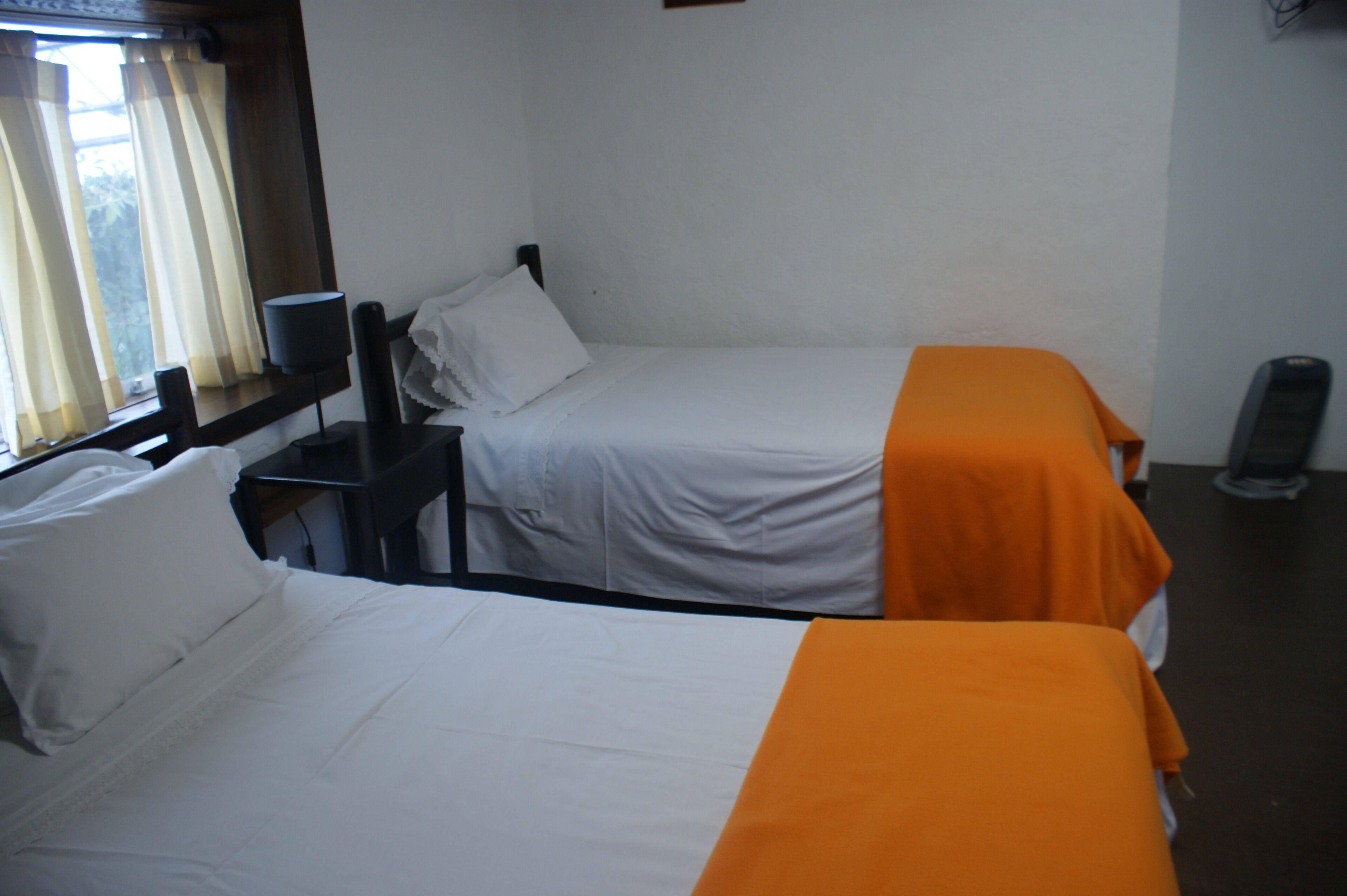 Una habitación con dos camas, una con cubrecama naranja y la otra con cubrecama blanca. Hay una mesita con una lámpara y un televisor encima.
