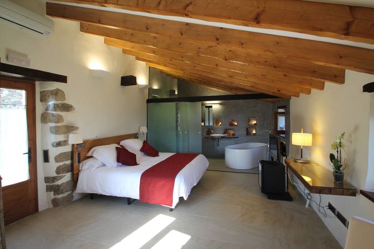 Quarto com cama, parede de pedra, teto de madeira, banheiro com banheira e pia.