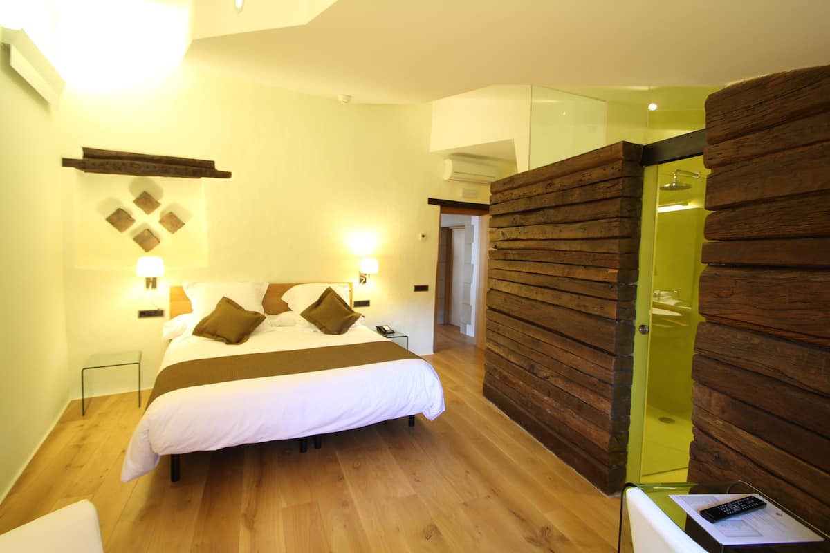 Quarto de hotel moderno com parede em madeira, uma cama grande com roupa de cama branca, uma mesa de cabeceira com abajur e uma pequena mesinha com controle remoto.