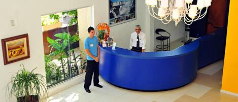 A reception desk with two staff members, a chandelier, framed pictures on the wall, and potted plants.