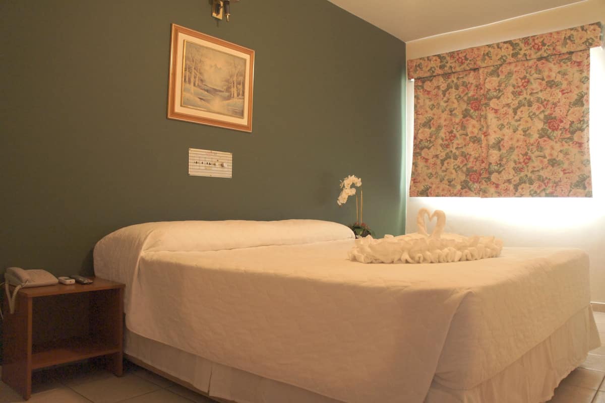A neatly made bed with a floral curtain, a framed picture on the wall, and a small bedside table with a phone and remote control.