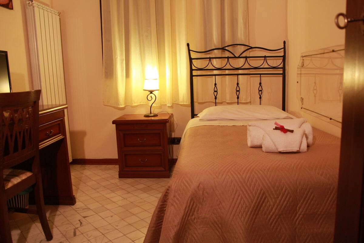 Una cama bien hecha con una colcha marrón, una mesita de noche con una lámpara, una silla de madera y un radiador.