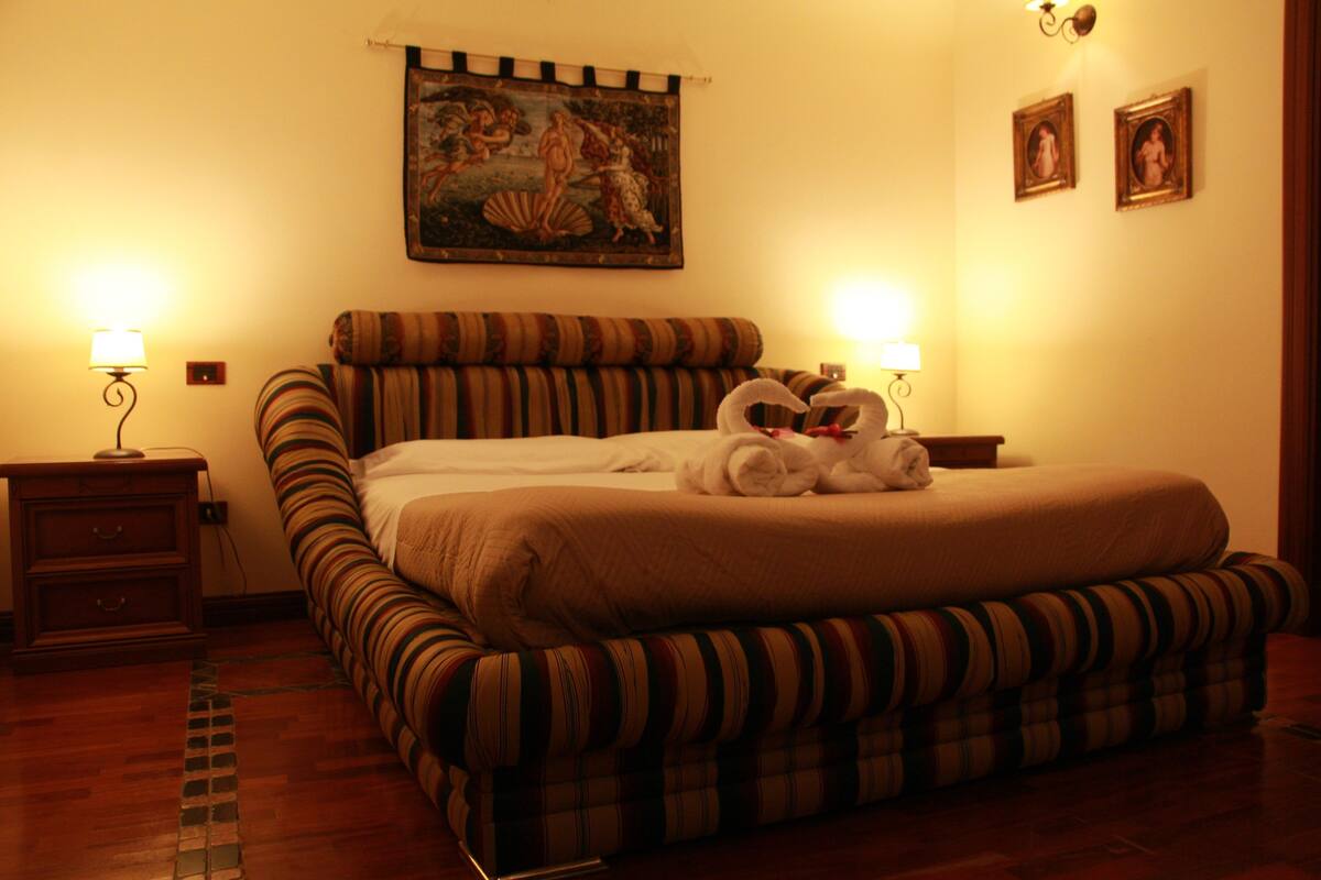 Una habitación de hotel con una cama grande, ropa de cama a rayas, dos lámparas de noche, un cuadro en la pared y una mesita de noche.