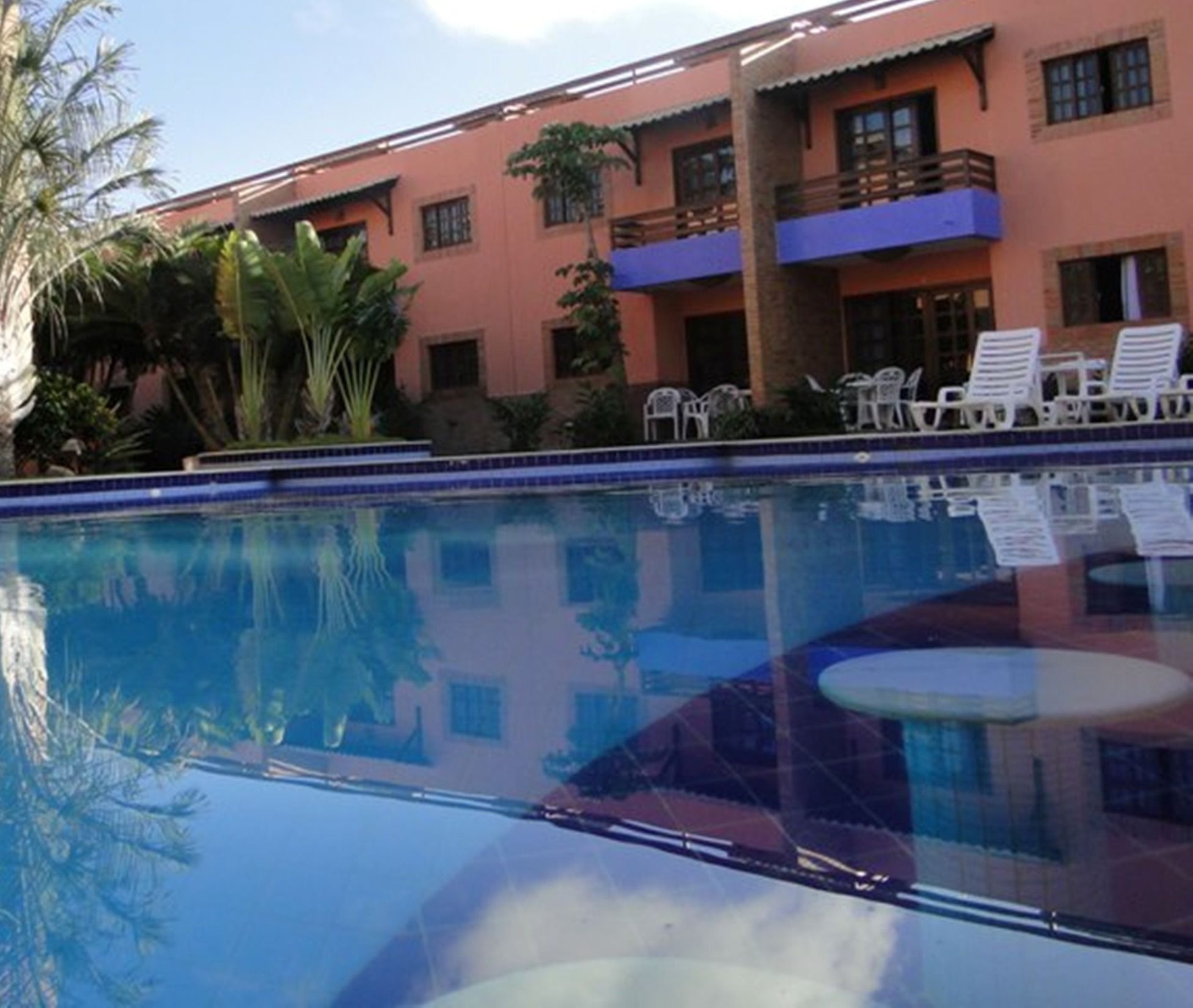 A swimming pool with a clear blue water surface, surrounded by white lounge chairs and a building with balconies.