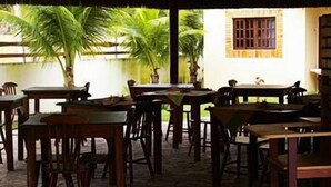 An indoor dining area with wooden tables and chairs, a thatched roof, and a view of palm trees outside. - CostaSol Pipa (Tibau do Sul)