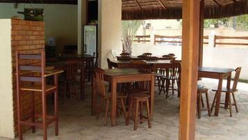 An outdoor dining area with wooden tables and chairs, a brick wall, and a thatched roof structure.