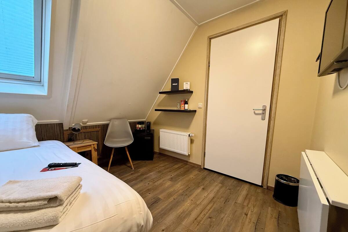 A hotel room with a bed, a chair, a desk, a radiator, a door, and a shelf with items.