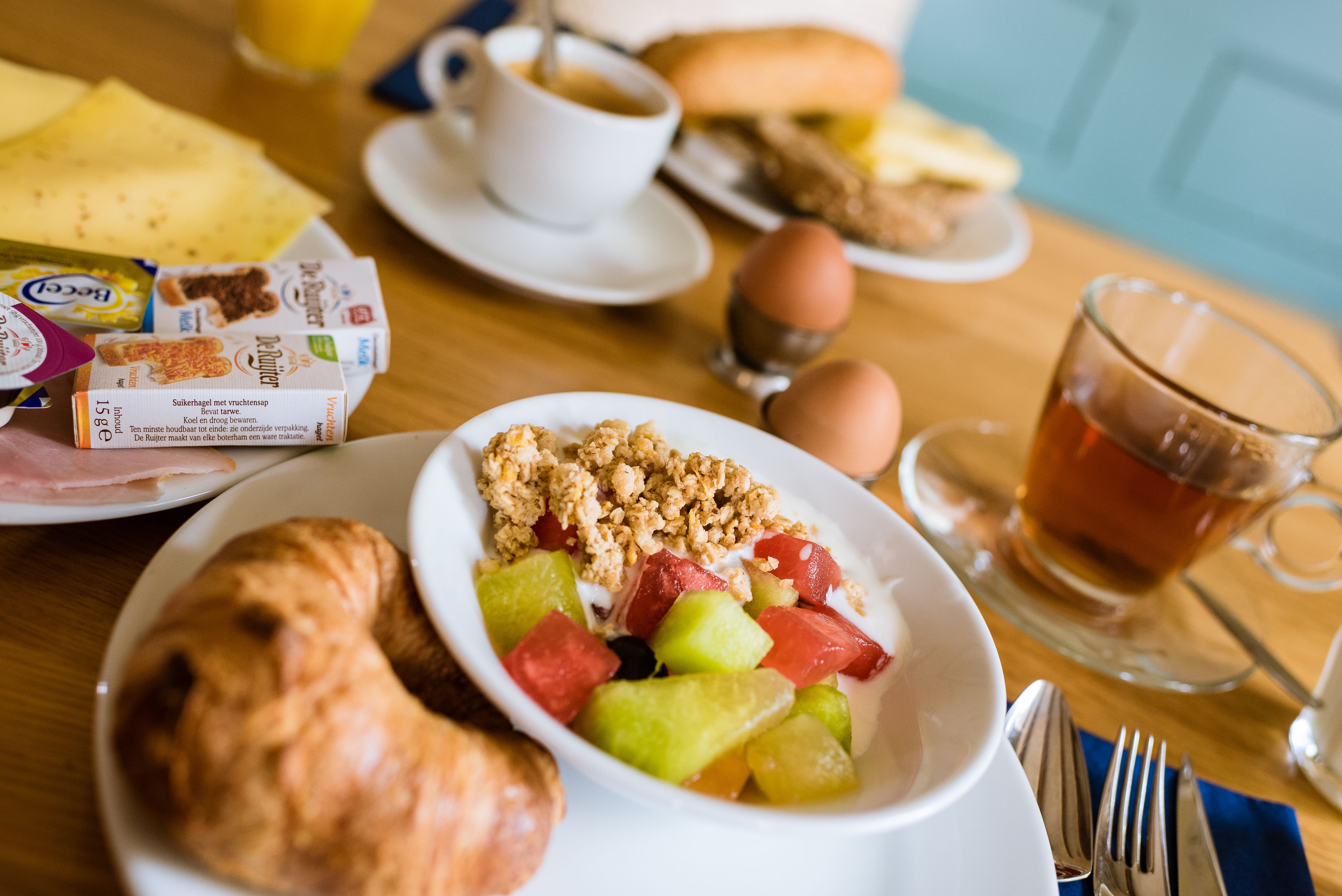 Een ontbijttafel met een bord fruit, yoghurt en granola, een croissant, een kop koffie en een glas thee.