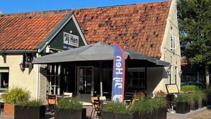 A building with a tiled roof, a sign reading "Bij Hen," outdoor seating with tables and chairs, and a flag. - Bij hen op Ameland (Hollum)