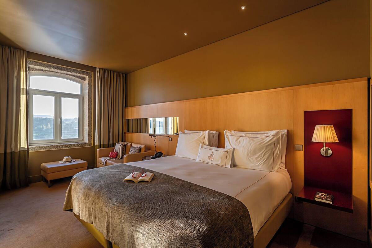Habitación de hotel con una cama grande, una mesita de noche, una lámpara y vista al mar a través de una ventana.