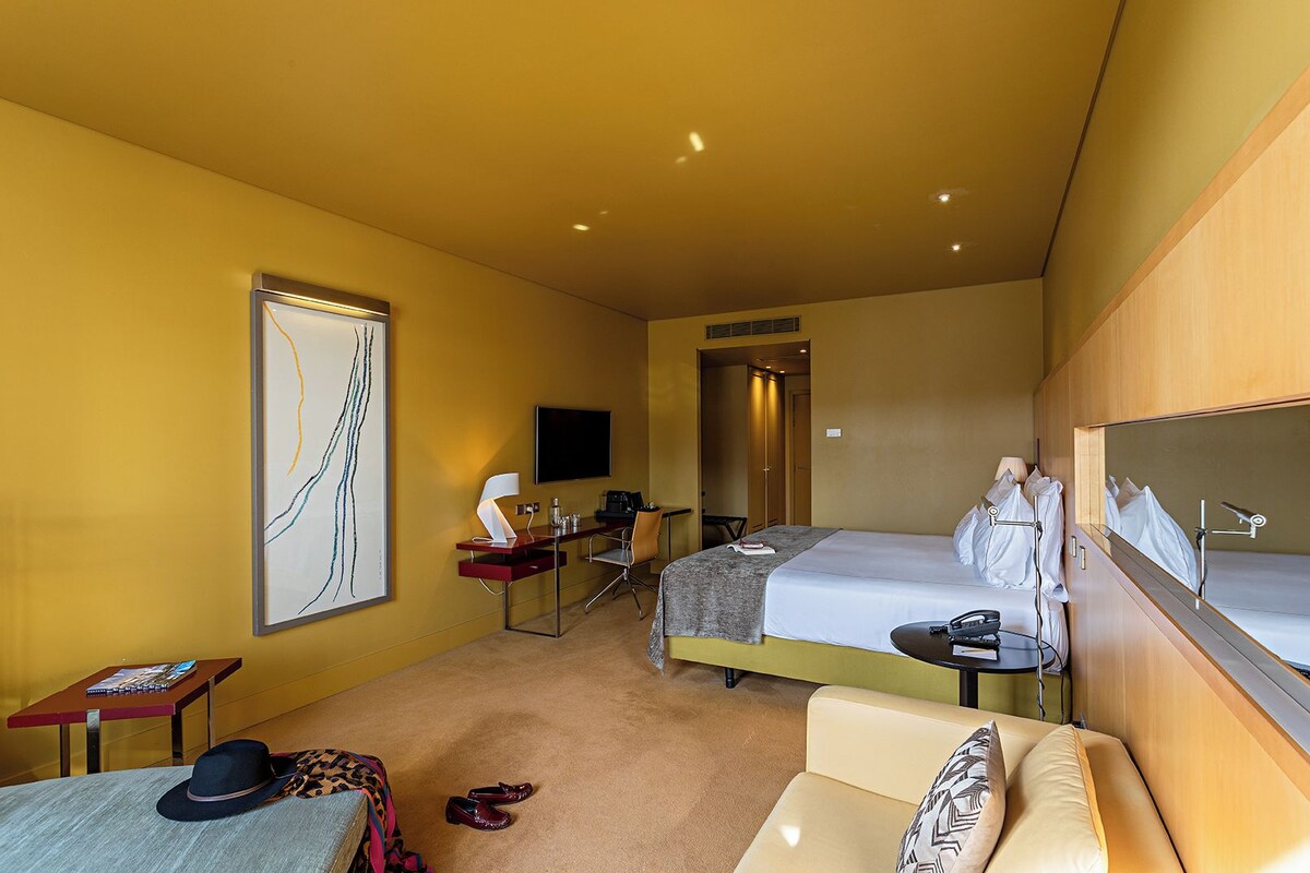 A hotel room with a bed, a desk, a chair, a sofa, a TV, and a large mirror.