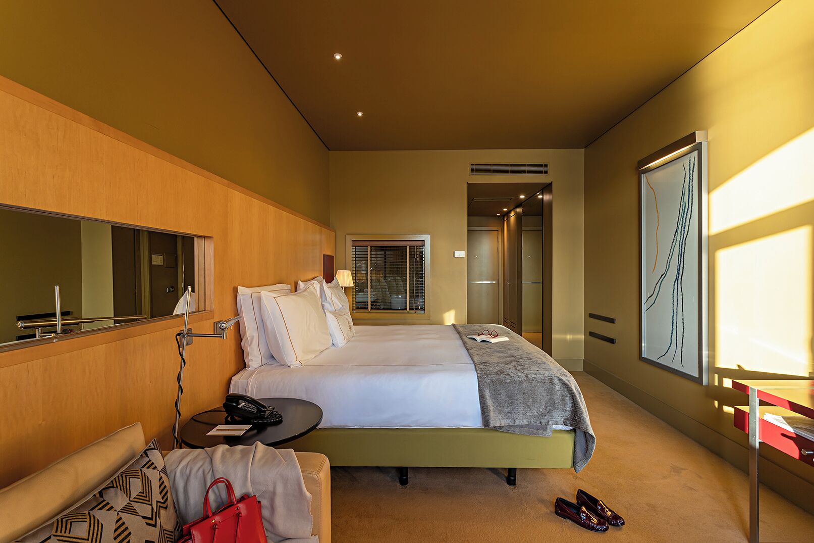 Habitación de hotel con cama, mesita de noche y espejo. Se ve una puerta que da a un baño y a un pasillo.