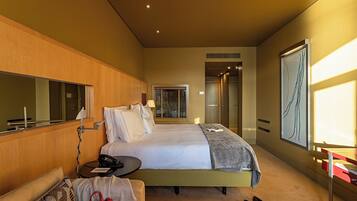 Habitación de hotel con cama, mesita de noche y espejo. Se ve una puerta que da a un baño y a un pasillo.