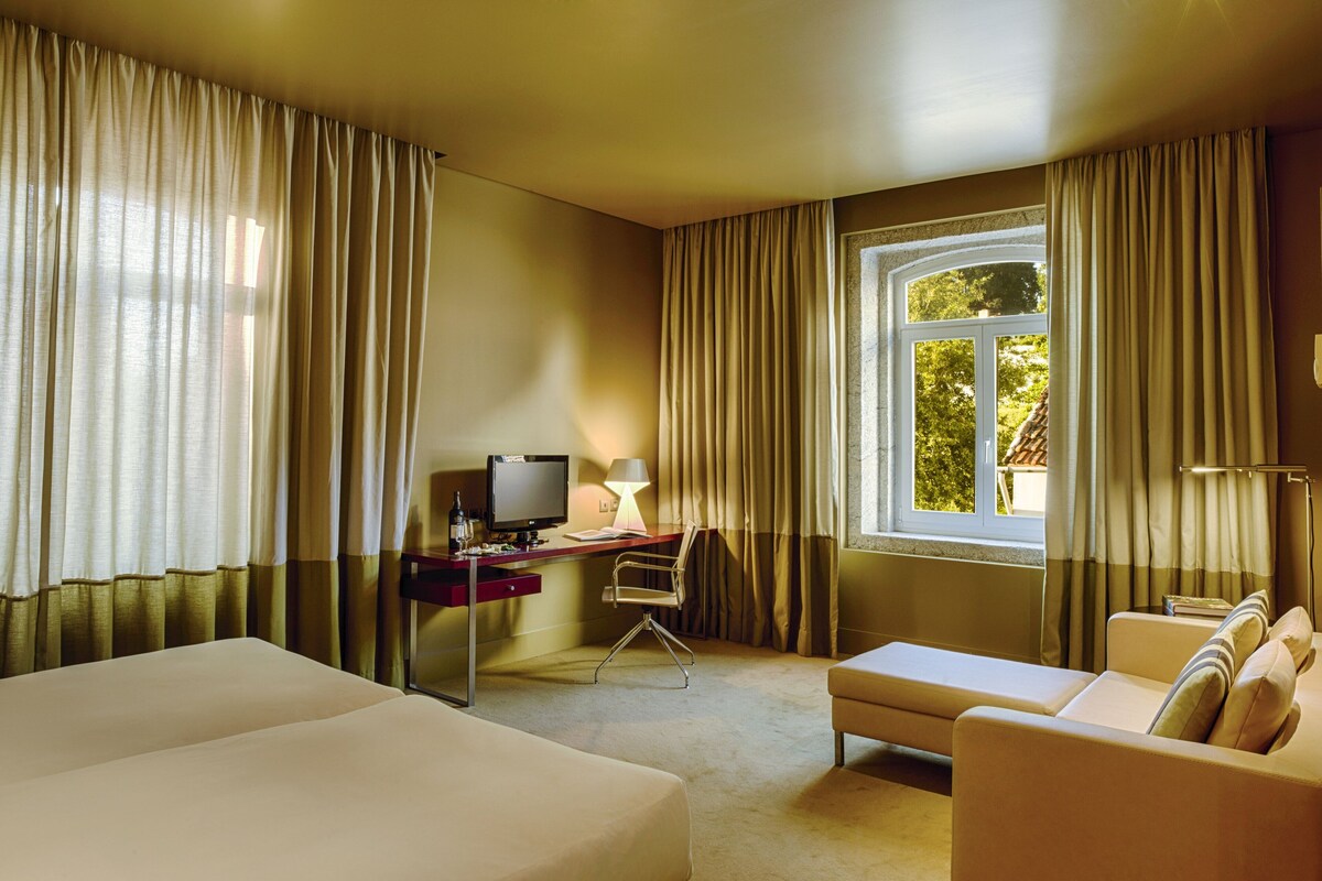 A hotel room with a large bed, a desk with a computer, a chair, a sofa, and a window with curtains.