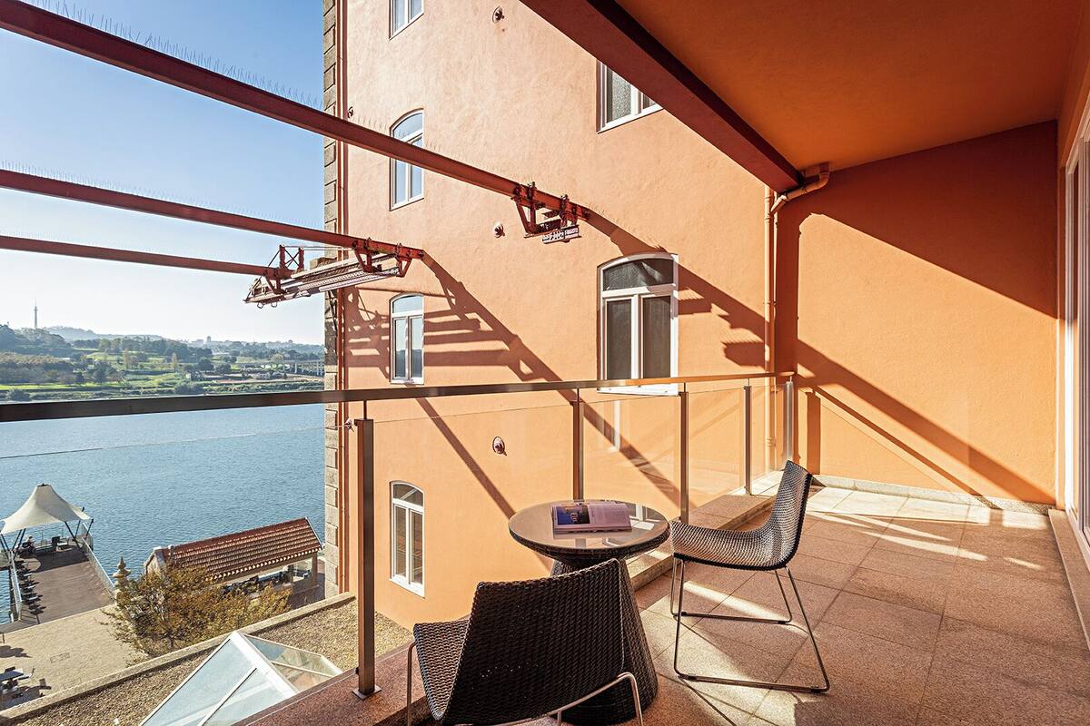 Un balcón con mobiliario de mimbre, una mesa y vistas a un río y un paisaje lejano.