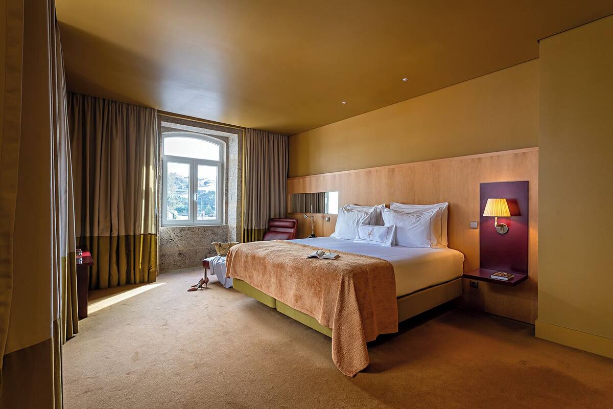 Habitación de hotel con cama, mesita de noche con lámpara, ventana con cortinas y vista al exterior.