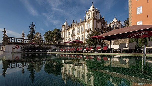 Piscina con el reflejo de un edificio histórico, sillas de descanso y una zona cubierta con sombrillas.
