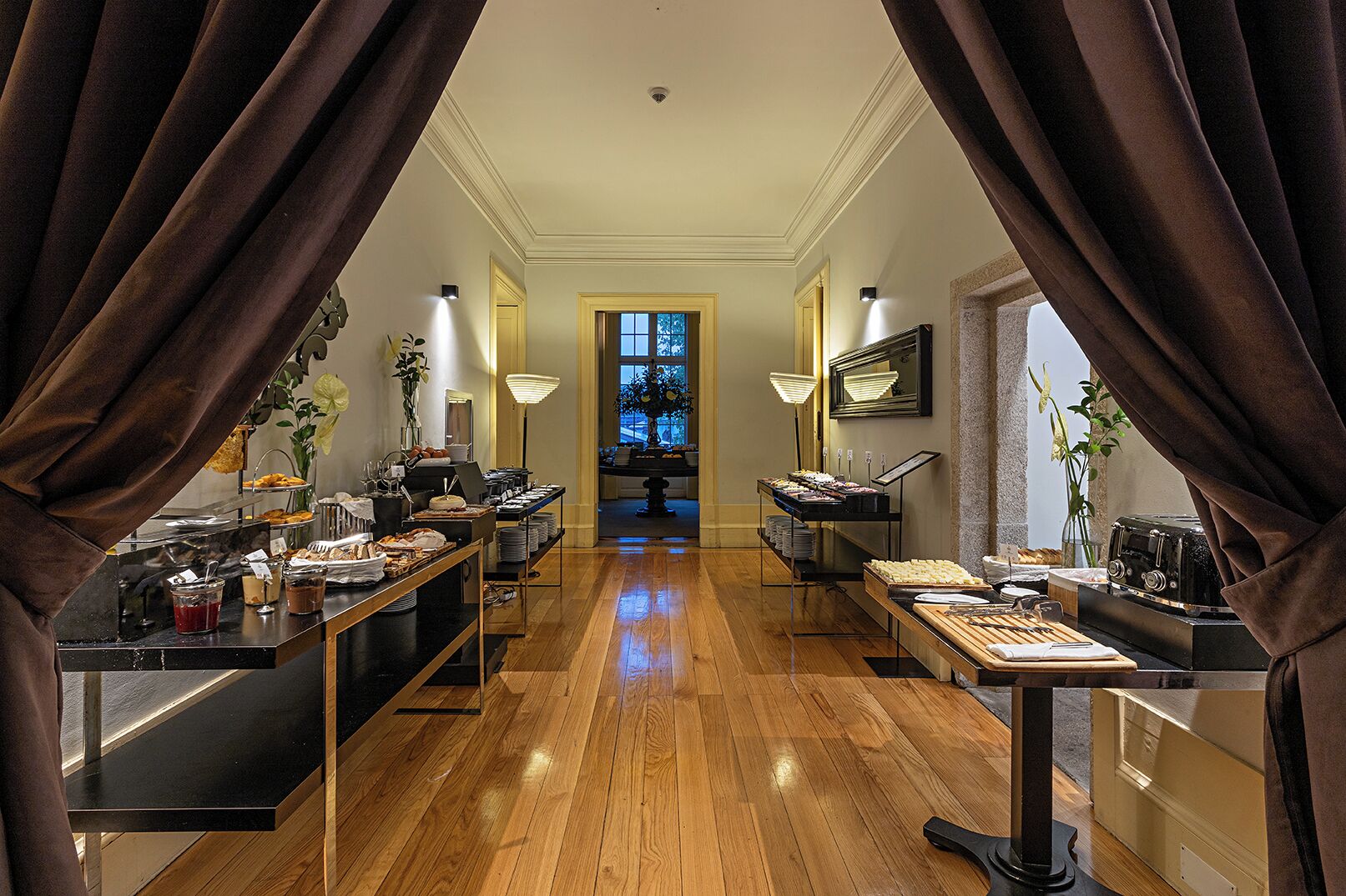 A hotel dining area with a buffet setup, wooden flooring, and a view into the dining room.