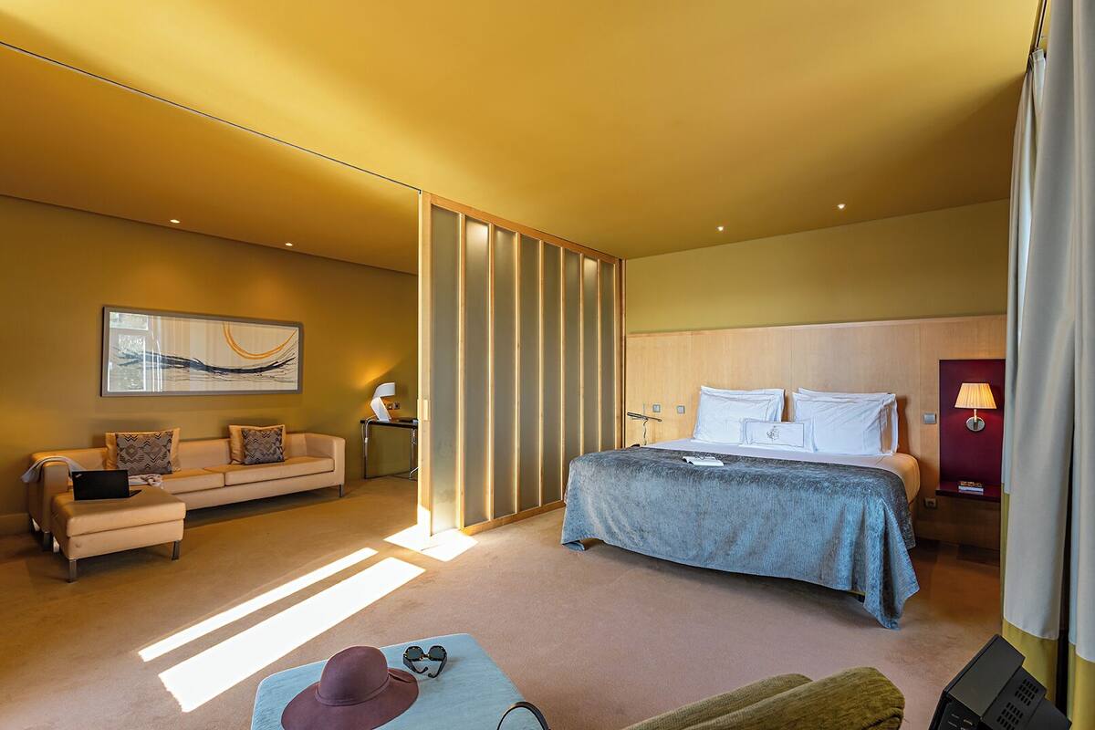 Habitación de hotel moderna con cama, sofá, escritorio y un amplio ventanal que deja entrar luz natural.